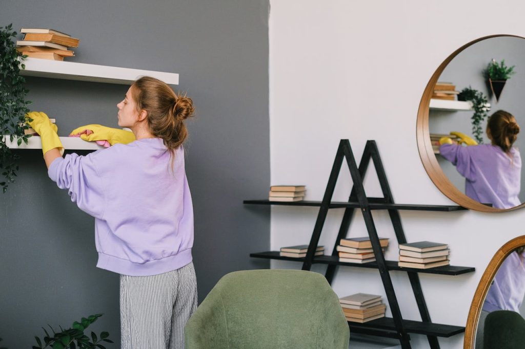 A woman cleans shelves in a stylish living room with mirrors and decor.